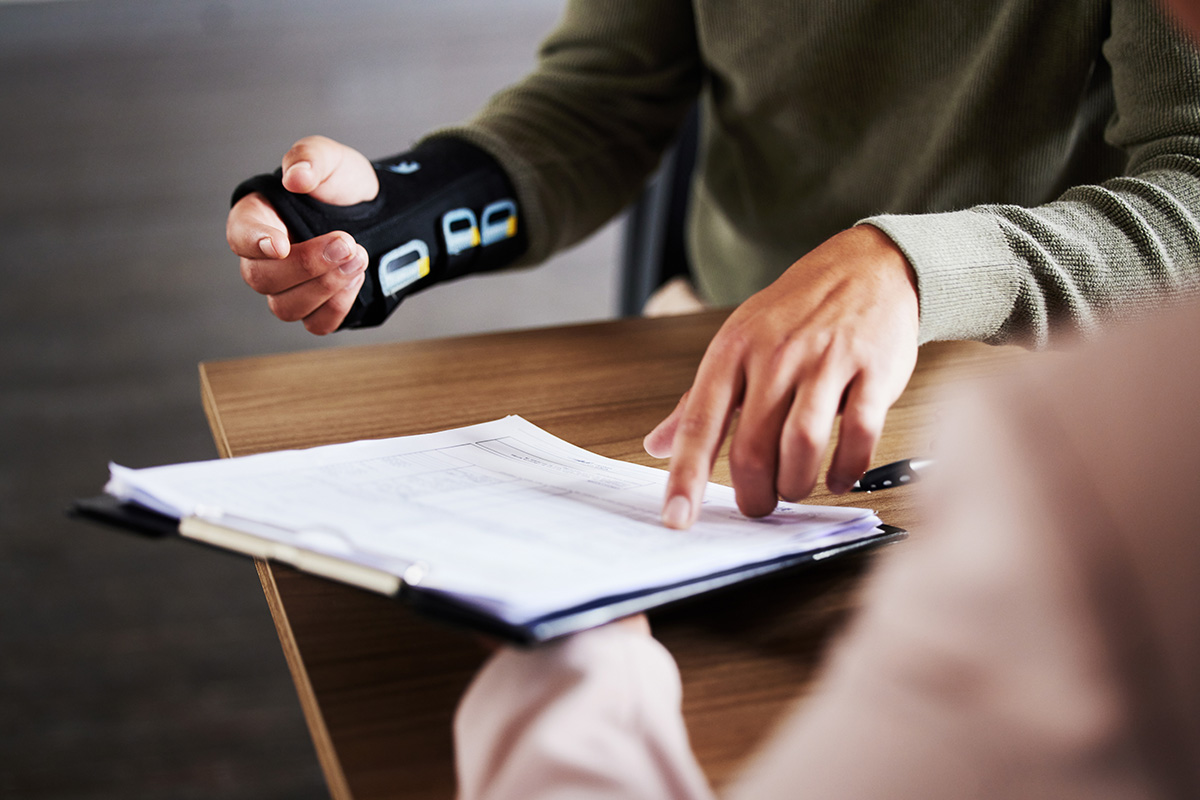 A person in a green sweater points at documents on a clipboard, while another with a wrist brace listens attentively.