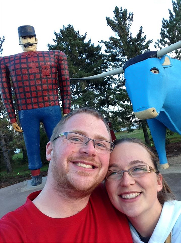 jordan-kretzschmar-sarah-bemidji Jordan and his wife, Sara takes a selfie in front of a large Paul Bunyan statue and a blue ox, with trees in the background.