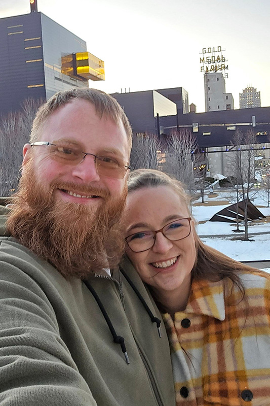 jordan-kretzschmar-sarah-minneapolis Jordan and his wife, Sara takes a selfie in front of modern buildings in Minneapolis at sunset. Jordan has a beard, and Sara wears glasses and a plaid coat.