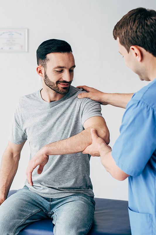 man-having-checkup A man in a gray shirt, sitting on a medical examination table, has his arm evaluated by a healthcare professional in blue scrubs.